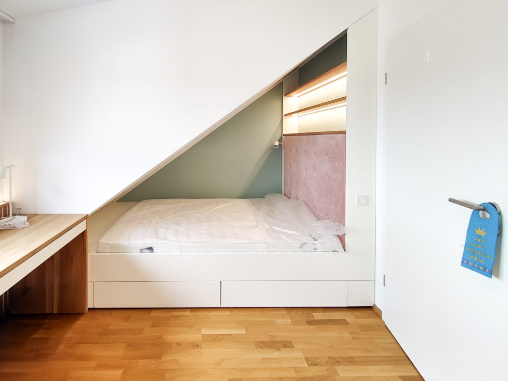 Nischenbett in der Dachschräge mit integriertem Stauraum und gepolstertem Kopfteil – maßgefertigte Einbaulösung im Kinderzimmer Neubau mit LED-Regal und Schreibtisch nach Maß.